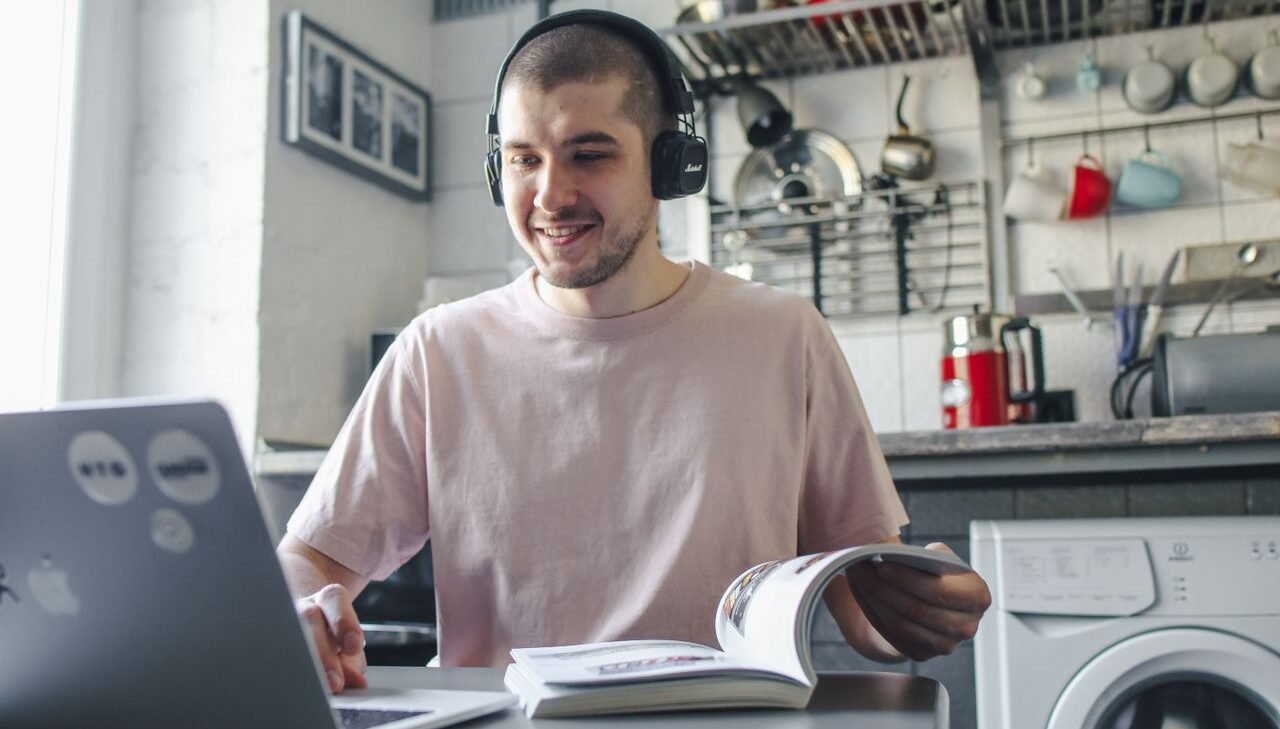 Smiling man wearing headphones, studying with a laptop and book in a cozy kitchen setting.